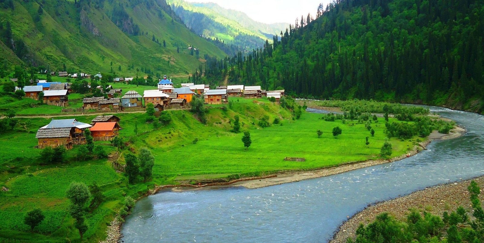 Scenic view of a lush green valley with wooden houses, a flowing river, and forested mountains in Azad Kashmir, Pakistan.